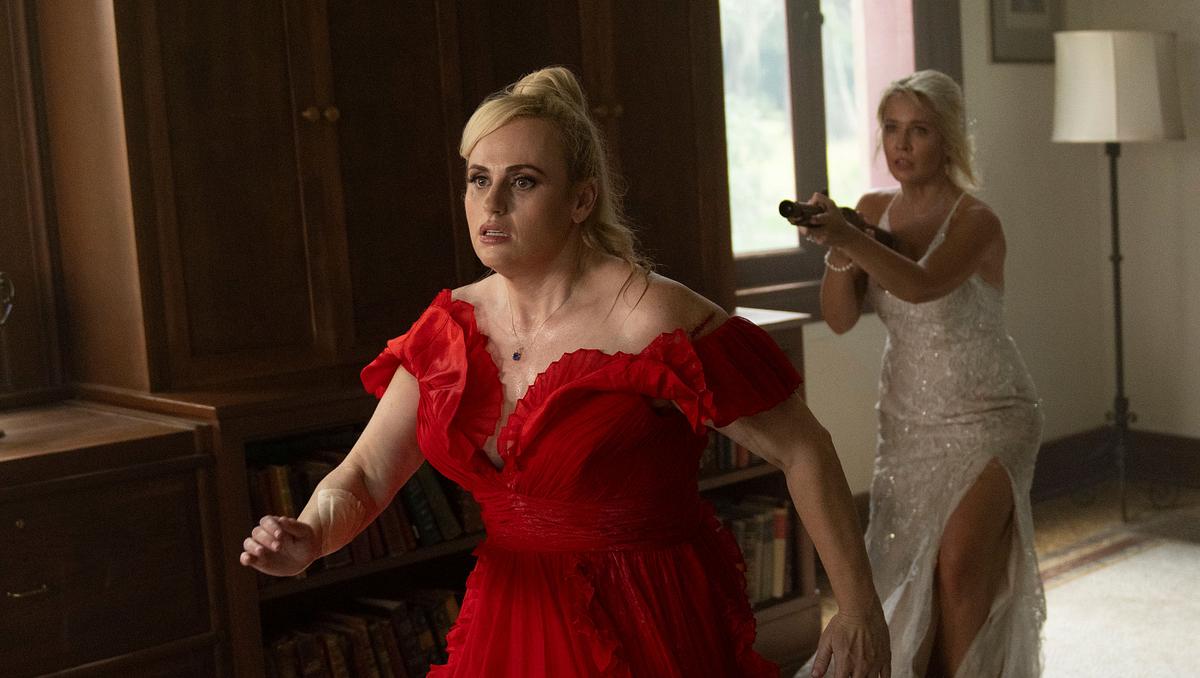 In einer Actionszene steht Rebel Wilson in rotem Kleid vorne, während Anna Camp im weißen Brautkleid mit einem Gewehr hinter ihr Deckung gibt.