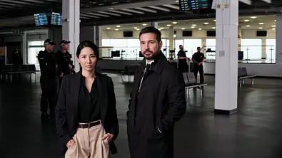 Hana Li und Clay Brody stehen in einem Flughafen-Terminal. Beide schauen ernst nach vorne. - Foto: Sony Pictures Entertainment. All Rights Reserved