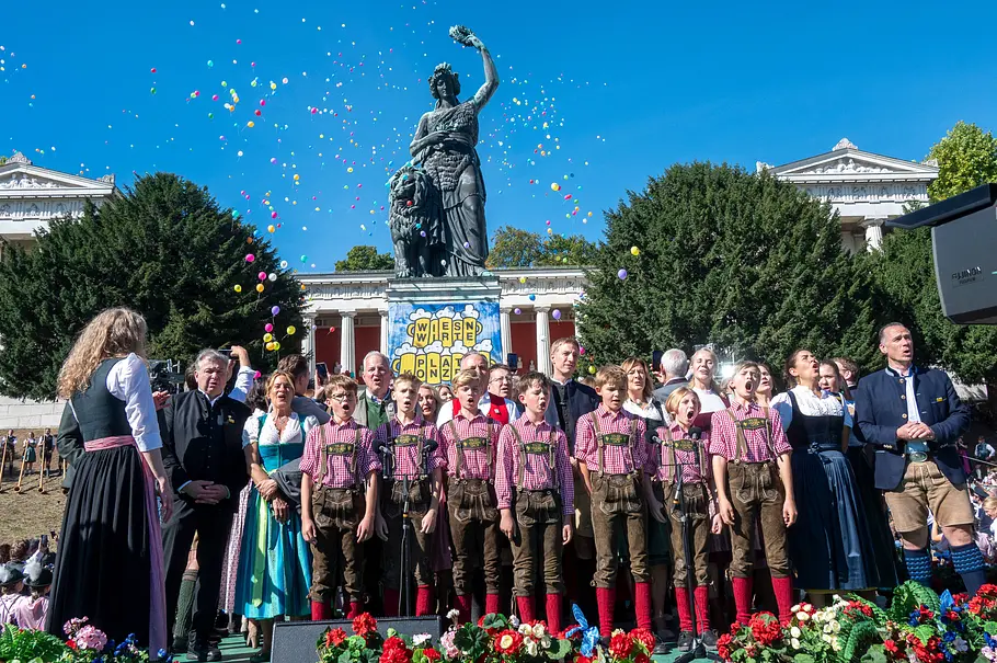 Regensburger Domspatzen ZDF Weihnachtsshow Die Regensburger Domspatzen singen mit Erwachsenen in Tracht vor einer Statue. Bunte Luftballons steigen in den Himmel.