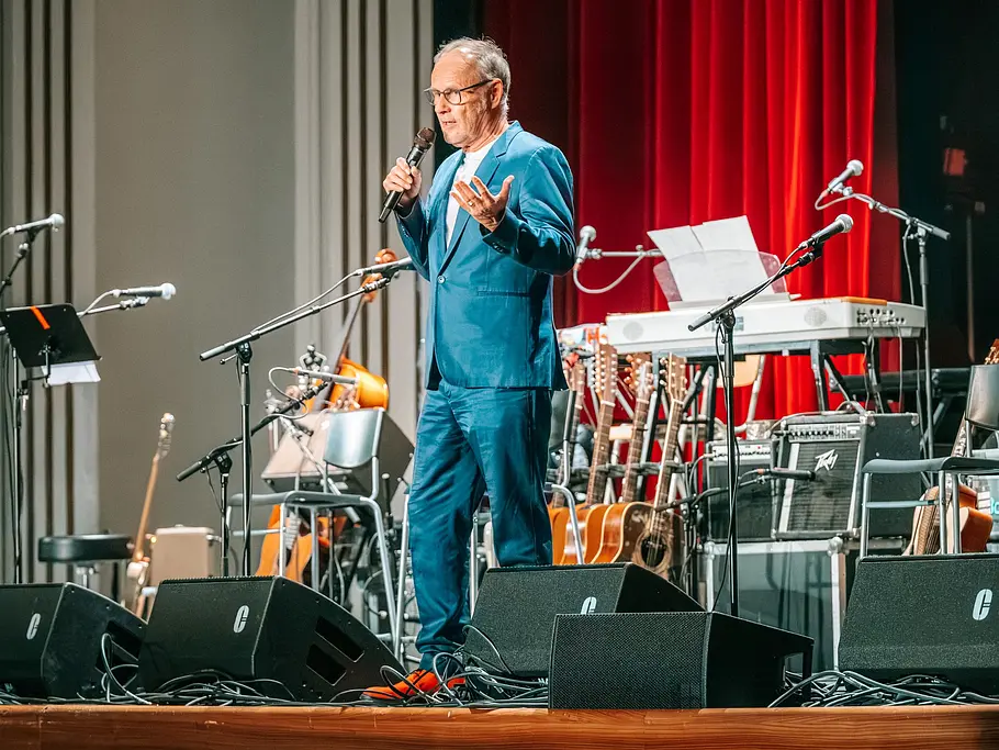 Reinhold Beckmann steht im blauen Anzug mit Mikrofon auf einer Bühne mit Instrumenten und Lautsprechern vor rotem Vorhang.