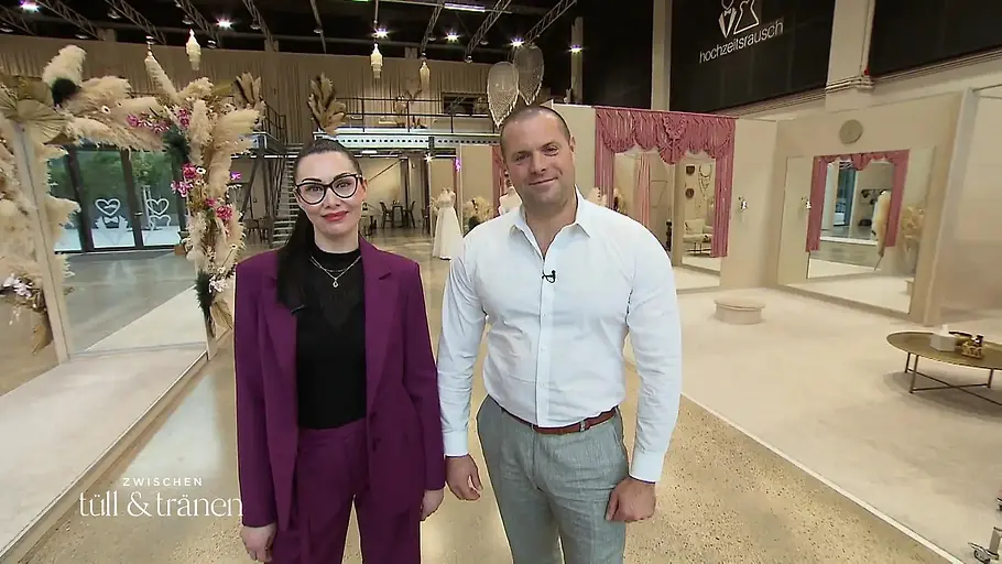 Rene Emser und Verlobte Sarah Zwei Personen stehen in einer hellen Messehalle vor dekorierten Ausstellungsständen und blicken in die Kamera.