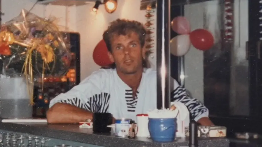 Redo lehnt hinter einer Theke mit Gläsern und Tassen. Neben ihm steht ein Blumenstrauß, im Hintergrund hängen Luftballons.