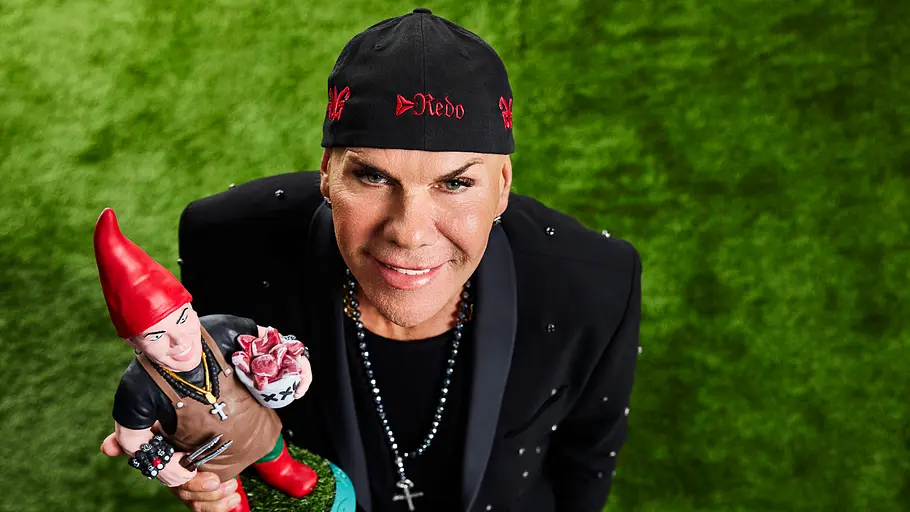 Rene Redo privat im Steckbrief Rene Redo trägt eine schwarze Kappe und hält einen Gartenzwerg mit roter Mütze. Er blickt auf grünem Rasen zur Kamera hinauf.