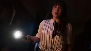 Eine junge Frau mit langen dunklen Haaren (Mia Tomlinson als Judy Warren) steht in einem dunklen Raum. Sie hält eine Taschenlampe, ihr Gesicht wirkt angespannt und erschrocken, während das Licht den Raum erhellt. - Foto: © 2025 Warner Bros. Entertainment Inc. All Rights Reserved.