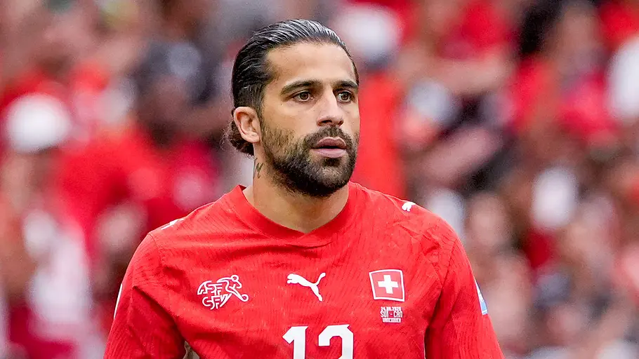 Ein Fußballspieler der Schweizer Nationalmannschaft im roten Trikot mit der Rückennummer 13 blickt während eines Spiels konzentriert nach vorne. Im Hintergrund ist die unscharfe Zuschauerkulisse des Stadions zu sehen.