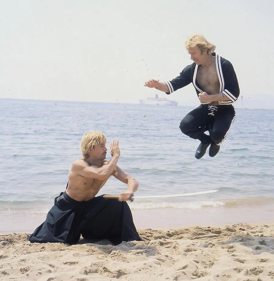 Richard Norton Chuck Norris. Zwei Männer üben Kampfkunst am Strand: Richard Norton kniet mit einem Schwert im Sand, Chuck Norris springt mit angezogenen Beinen.