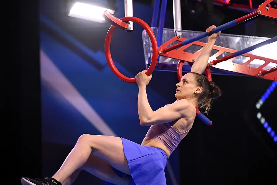 Rita Benker ist Teilnehmerin bei Ninja Warrior Germany 2026 Rita Benker hängt mit angewinkelten Beinen an einem roten Ring und einer blauen Stange eines Hindernisparcours.