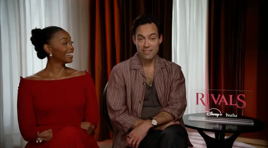 Nafessa Williams und Alex Hassell Nafessa Williams und Alex Hassell sitzen in einem Junket-Setting für „Rivals“. Sie lacht in seine Richtung, während er nach vorne schaut.
