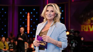 Kim Fisher steht in einem TV-Studio, hält eine ‚Riverboat‘-Karte in der Hand und lächelt; im Hintergrund sind Publikum und Studioleuchten zu sehen. - Foto: IMAGO / STAR-MEDIA