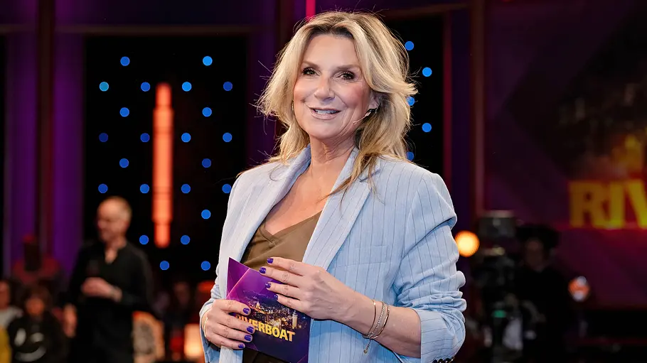 Kim Fisher Kim Fisher steht in einem TV-Studio, hält eine ‚Riverboat‘-Karte in der Hand und lächelt; im Hintergrund sind Publikum und Studioleuchten zu sehen.