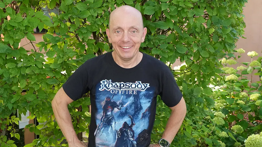 Bernhard Hoëcker Glatzköpfiger Mann in schwarzem Band‑T-Shirt steht lächelnd mit den Händen in die Hüften gestützt vor einem grünen Strauch im Garten.