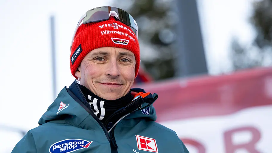 Eric Frenzel Mann in grüner Winterjacke mit Sponsorenlogos und roter Strickmütze steht im Freien vor unscharfem Hintergrund einer Wintersportanlage.