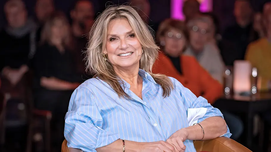Riverboat Blondhaarige Moderatorin sitzt lächelnd in einem Ledersessel in einem TV-Studio, im Hintergrund unscharfes Publikum und stimmungsvolle Beleuchtung.