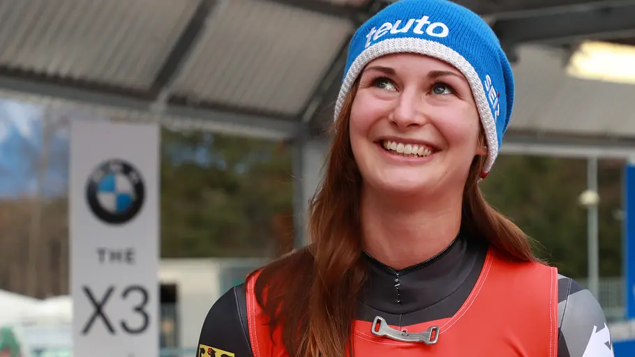 Julia Taubitz Lächelnde Sportlerin in Rennanzug und blauer Mütze steht in einer Wintersportanlage vor Werbebanden