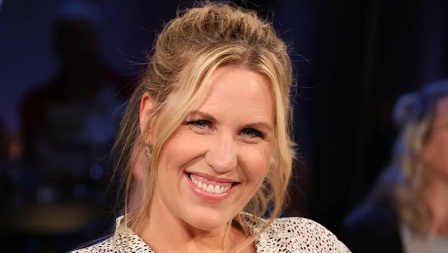 Nicole Staudinger Blondhaarige Frau mit hochgesteckten Haaren lächelt in einem Studio mit dunklem, unscharfem Hintergrund.