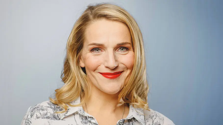 Tanja Wedhorn Blondhaarige Frau mit rotem Lippenstift lächelt vor neutralem Hintergrund in die Kamera.