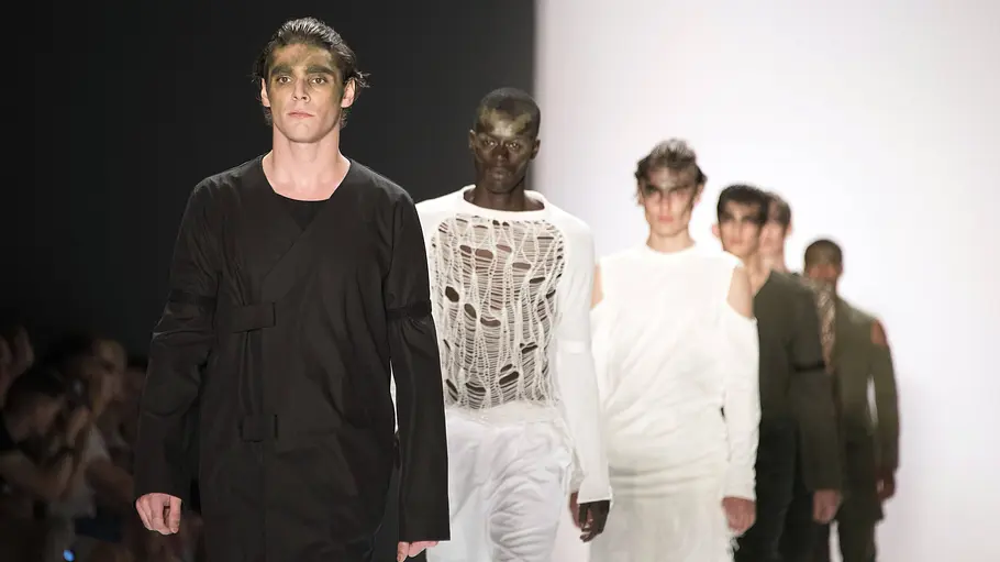 RJ Mitte führt in einem schwarzen Outfit eine Reihe von Models in weißen und dunklen Entwürfen über den Laufsteg.