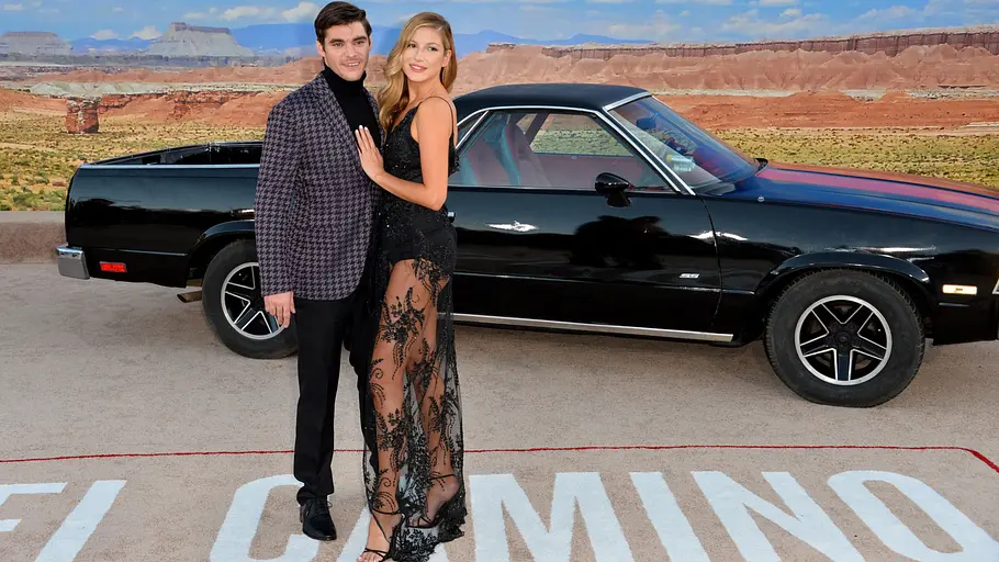 RJ Mitte im gemusterten Sakko und eine Frau im schwarzen Spitzenkleid posieren vor einem schwarzen Auto und einer Wüstenkulisse.