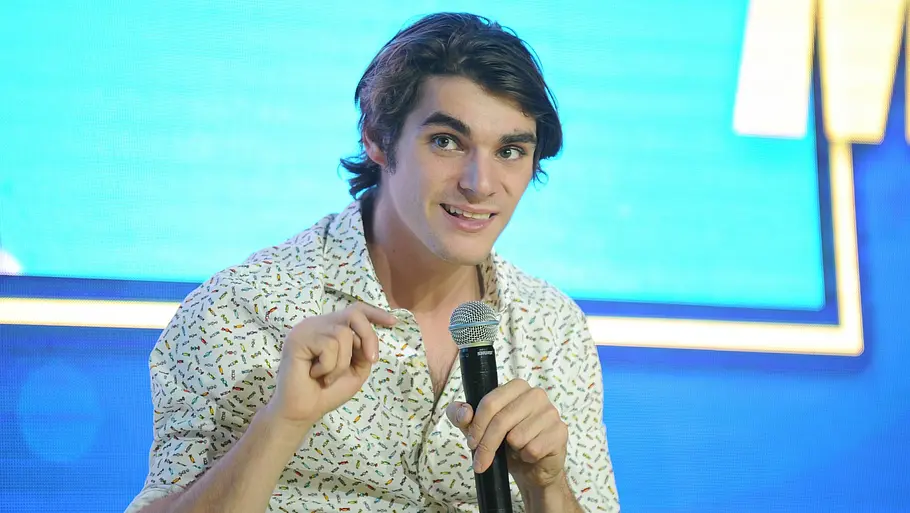 RJ Mitte sitzt in einem hellen, gemusterten Hemd vor blauem Hintergrund, hält ein Mikrofon und gestikuliert mit der freien Hand.