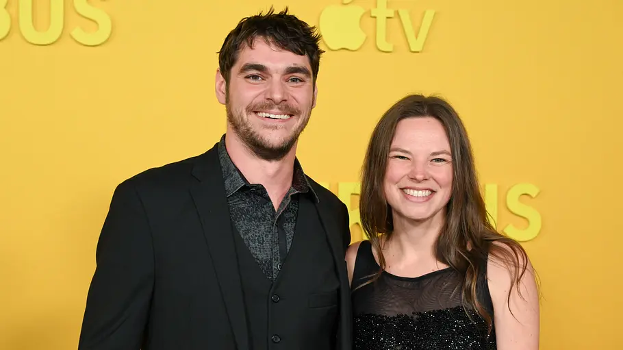 RJ Mitte im schwarzen Anzug und Kennedy Suarez im schwarzen Glitzerkleid lächeln vor einer gelben Wand mit Apple-TV-Logo.