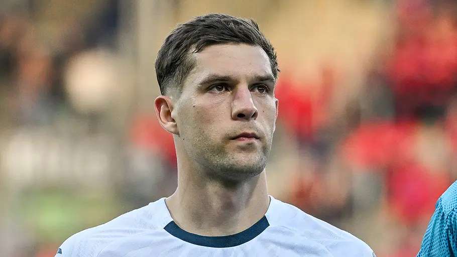Ein Fußballspieler der TSG Hoffenheim steht im weißen Trikot mit ernstem Blick vor dem Anpfiff auf dem Spielfeld. Im Hintergrund sind die Zuschauer im Stadion unscharf zu erkennen.
