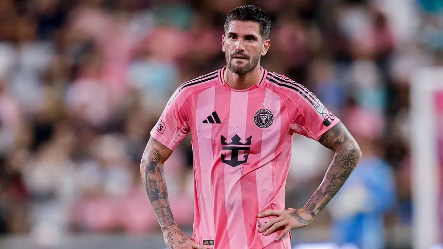 Ein Fußballspieler von Inter Miami im pinken Trikot mit der Rückennummer 7 steht während eines MLS-Spiels mit den Händen in den Hüften auf dem Rasen. Im Hintergrund sind Zuschauer und das Spielfeld unscharf zu erkennen.