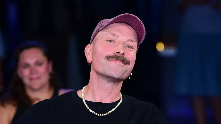 Roger G. Influencer Roger G. lächelt während einer TV-Aufzeichnung in die Kamera und trägt eine rosafarbene Cap sowie ein schwarzes T-Shirt.