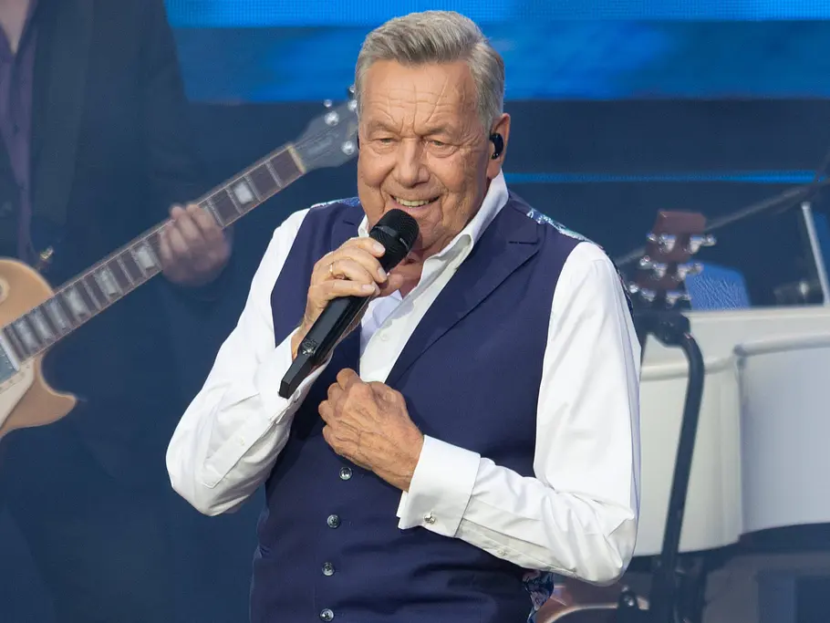 Roland Kaiser bei der „Helene Fischer Show“ 2026 Roland Kaiser singt im weißen Hemd und blauer Weste ins Mikrofon. Hinter ihm stehen ein Gitarrist und ein weißer Flügel.