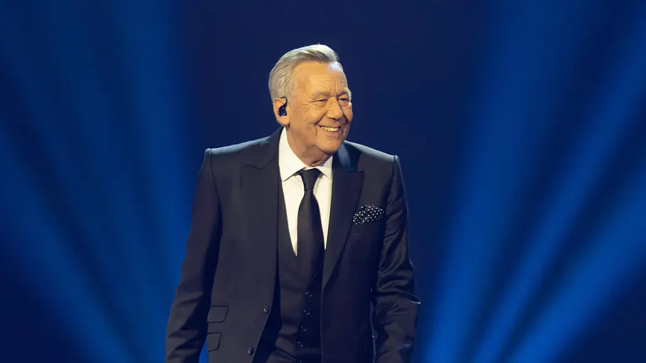 Roland Kaiser Roland Kaiser trägt einen schwarzen Anzug. Er steht auf der Bühne und lächelt.