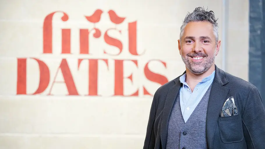 Roland Trettl First Dates Roland Trettl steht lächelnd in dunklem Sakko, grauer Weste und hellblauem Hemd vor dem roten Schriftzug „first DATES“.