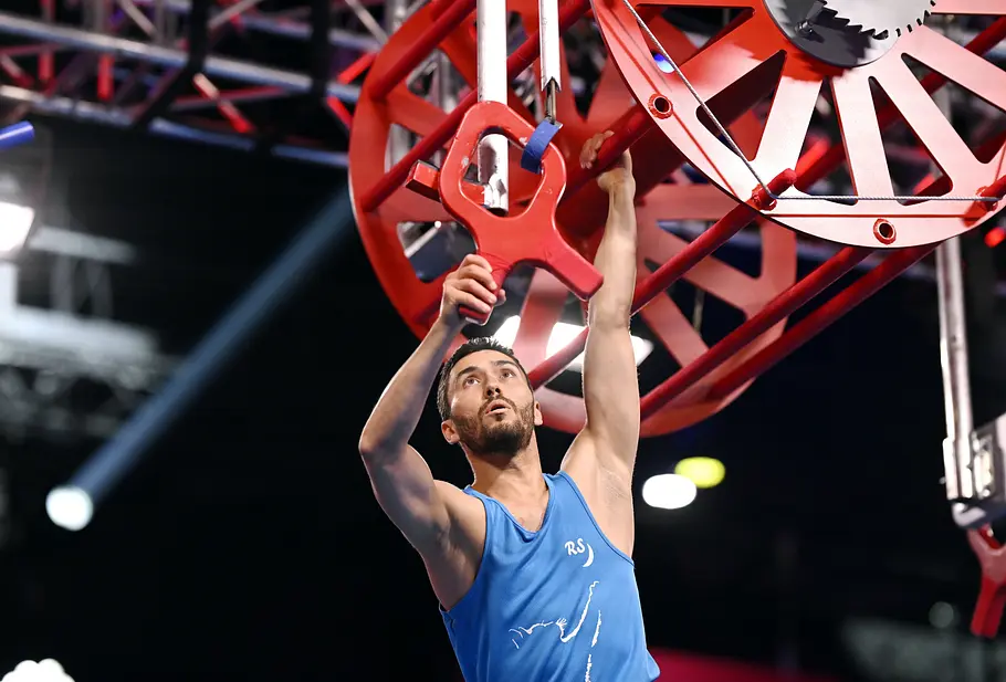 Roman Schirillef ist Teilnehmer bei Ninja Warrior Germany 2026 Roman Schirillef hält sich im blauen Sportshirt mit erhobenen Armen an einem roten Hindernis fest.
