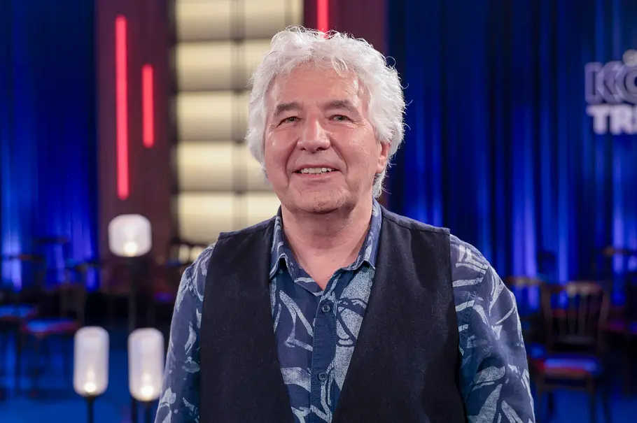 Ronald Schnabel Ronald Schnabel lächelt mit weißem Haar, blau gemustertem Hemd und dunkler Weste vor blau beleuchteten Vorhängen.