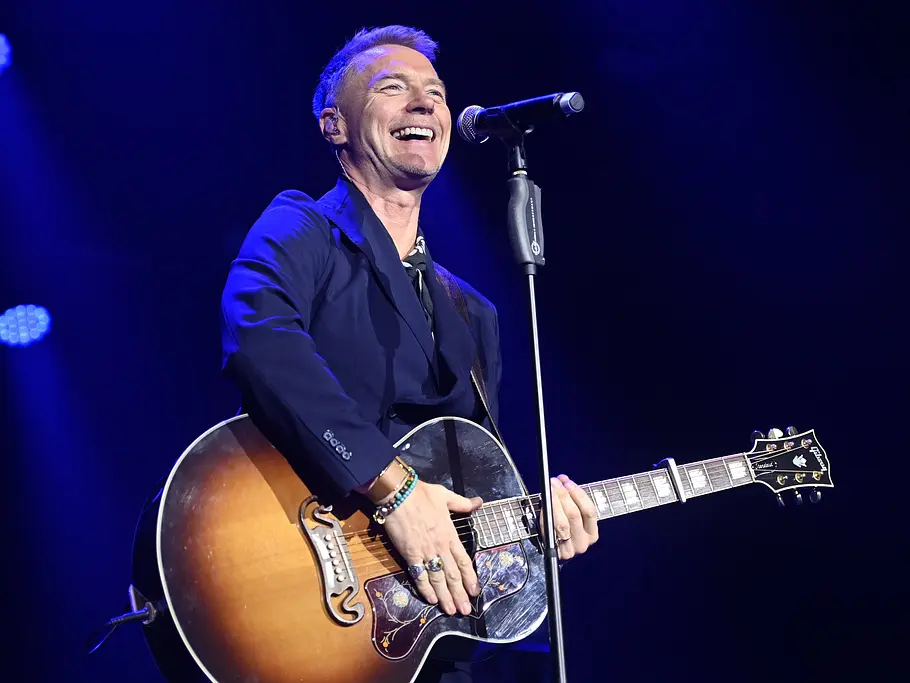 Ronan Keating steht lächelnd mit einer Akustikgitarre vor einem Mikrofon auf einer blau beleuchteten Bühne.