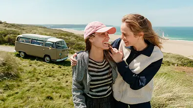 „Rosamunde Pilcher: Wo Liebe aufhört und wo sie beginnt“: Auf einem grünen, hügeligen Küstenabschnitt oberhalb eines Sandstrands: Ivy Stevens (Rocío Luz) und Finn Cumberwell (Athena Strates) stehen nebeneinander und halten sich freundschaftlich im Arm. Beide blicken sich an und lachen. Im Hintergrund steht ein beigefarbener Oldtimer-VW-Bus. - Foto: ZDF/Steve Tanner