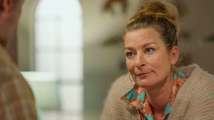„Rote Rosen“ Folge 4395, 9. Oktober 2026 Rote Rosen-Vorschau, Folge 4395: Carla trägt einen beigen Strickpullover über einer bunt gemusterten Bluse und schaut Michael an, der links unscharf zu sehen ist.