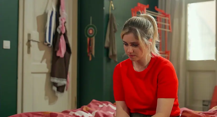 Rote Rosen Toni Toni sitzt mit gesenktem Blick und zusammengebundenen blonden Haaren im roten Oberteil auf einem Bett mit gemusterter Decke.