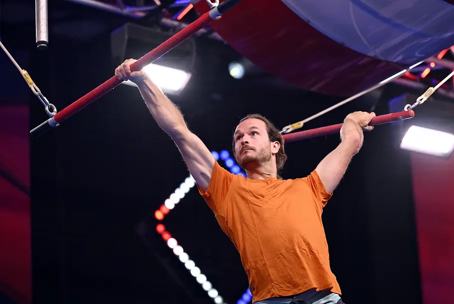 Ruben Kälin ist Teilnehmer bei Ninja Warrior Germany 2026 Ruben Kälin hängt im orangefarbenen T-Shirt mit weit gespreizten Armen an zwei roten Stangen eines Hindernisparcours.