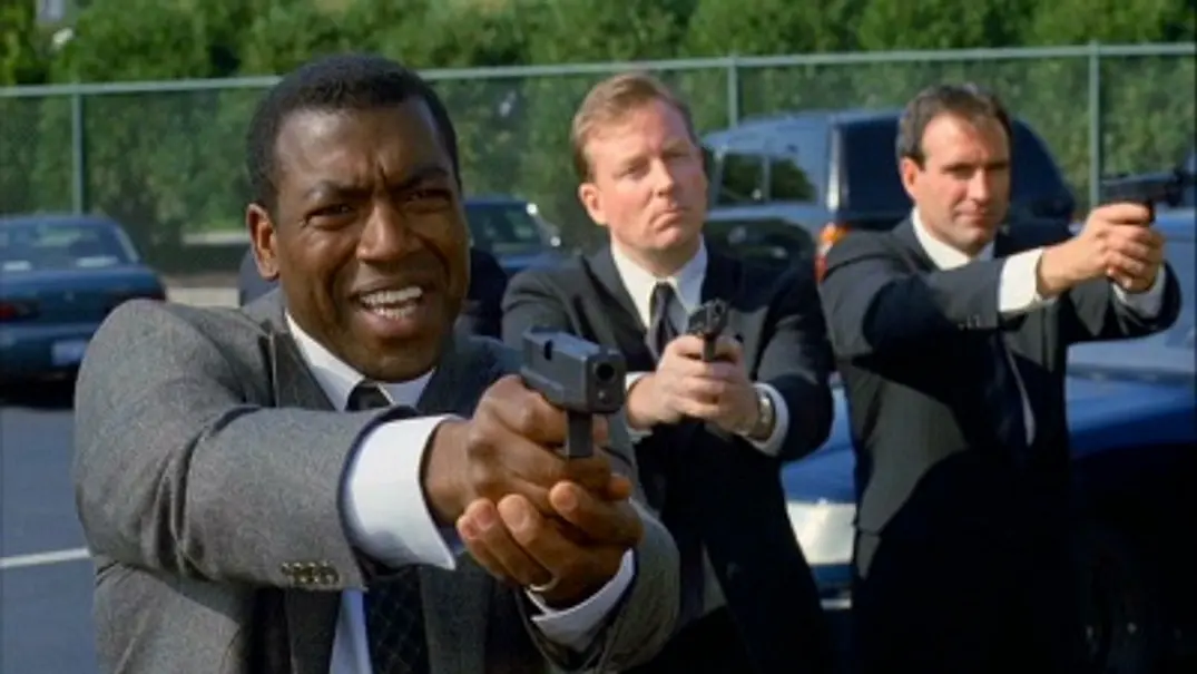 Russell Andrews zielt für den Film „Ein ungleiches Paar“ mit einer Waffe. Hinter ihm stehen zwei weitere Personen, die dasselbe tun. - Foto: © Warner Bros.