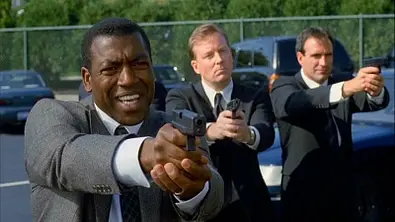 Russell Andrews zielt für den Film „Ein ungleiches Paar“ mit einer Waffe. Hinter ihm stehen zwei weitere Personen, die dasselbe tun. - Foto: © Warner Bros.