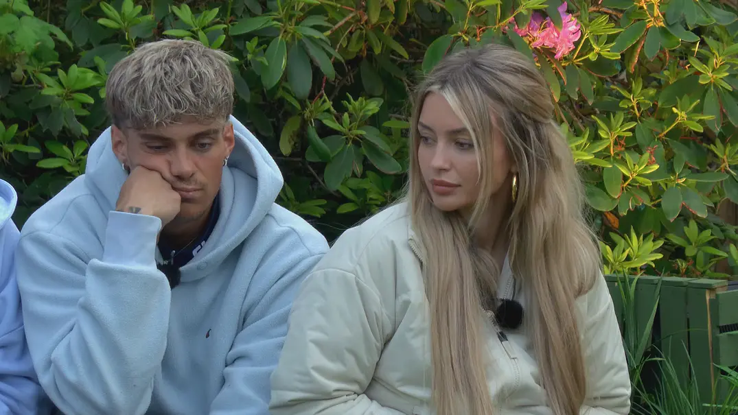 Ryan und Lina im Sommerhaus der Stars sitzen nebeneinander im Freien vor dichtem, grünem Buschwerk mit rosa Blüten.  - Foto: RTL