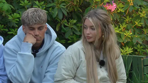 Ryan und Lina im Sommerhaus der Stars sitzen nebeneinander im Freien vor dichtem, grünem Buschwerk mit rosa Blüten.  - Foto: RTL