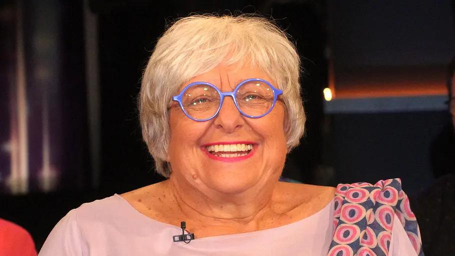 Sabine Asgodom Eine lächelnde Frau mit kurzen weißen Haaren und runder blauer Brille trägt eine hellrosa Bluse und ein gemustertes Tuch.