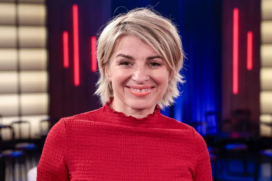 Sabine Heinrich Sabine Heinrich mit kurzen blonden Haaren und rotem Oberteil lächelt vor einem blau und rot beleuchteten Hintergrund.