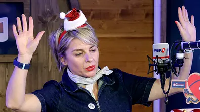 Moderatorin Sabine Heinrich trägt eine Weihnachtsmütze und hebt die Hände feierlich hoch. - Foto: © WDR/Thomas Brill
