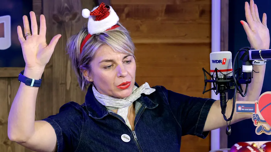 Sabine Heinrich mit Weihnachtsmütze. Moderatorin Sabine Heinrich trägt eine Weihnachtsmütze und hebt die Hände feierlich hoch.