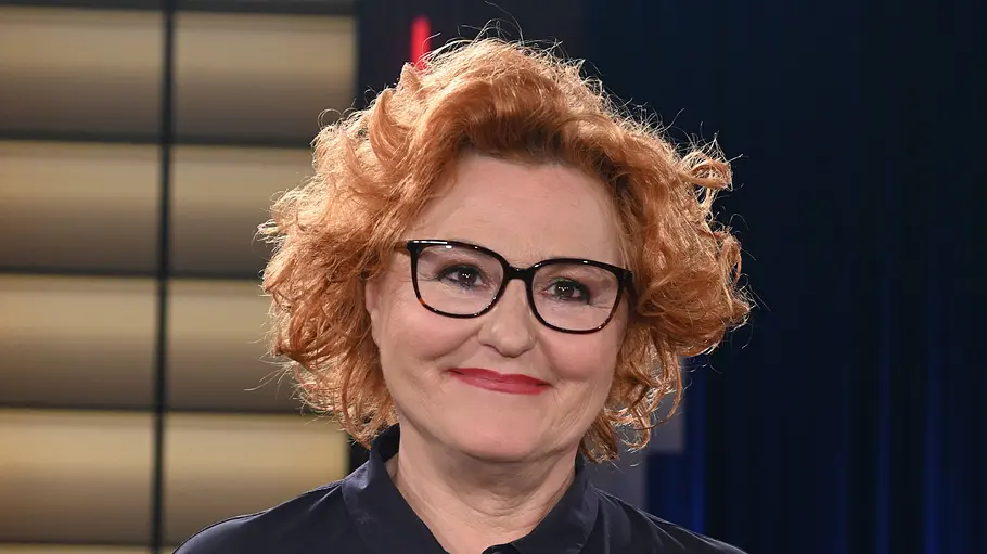 Sabine Rückert Sabine Rückert lächelt leicht in die Kamera. Sie trägt eine dunkle Bluse. Im Hintergrund ist ein TV‑Studio mit warm beleuchteten Wandelementen und dunklem Vorhang zu sehen.