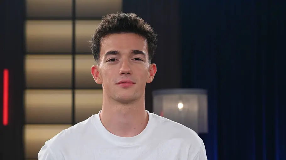 Sacha Wenk Sacha Wenk steht vor der Kamera und blickt neutral nach vorn. Er trägt ein weißes T‑Shirt. Im Hintergrund ist ein TV‑Studio mit dunklen Vorhängen, einem beleuchteten Wandpanel und roten vertikalen Lichtstreifen zu sehen.