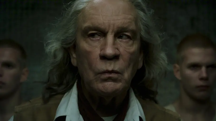 „Sacrifice“: John Malkovich übernimmt die Rolle des Gunnar John Malkovich mit langen grauen Haaren blickt ernst zur Seite. Hinter ihm stehen zwei unscharf erkennbare Männer im Dunkeln.