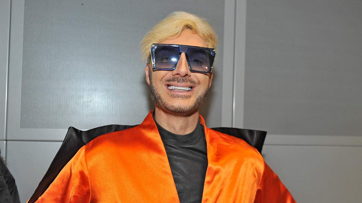 Sam Dylan lächelt in einem orangen Outfit und Sonnenbrille in die Kamera.