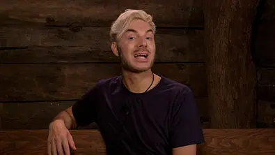 Sam Dylan sitzt in einer Holzhütte und spricht. Er trägt ein dunkles T-Shirt und hat blond gefärbte Haare - Foto:  RTL (DE)